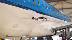 A black drone hovering next to a white and blue airplane inside a hangar A black drone hovering next to a white and blue airplane inside a hangar