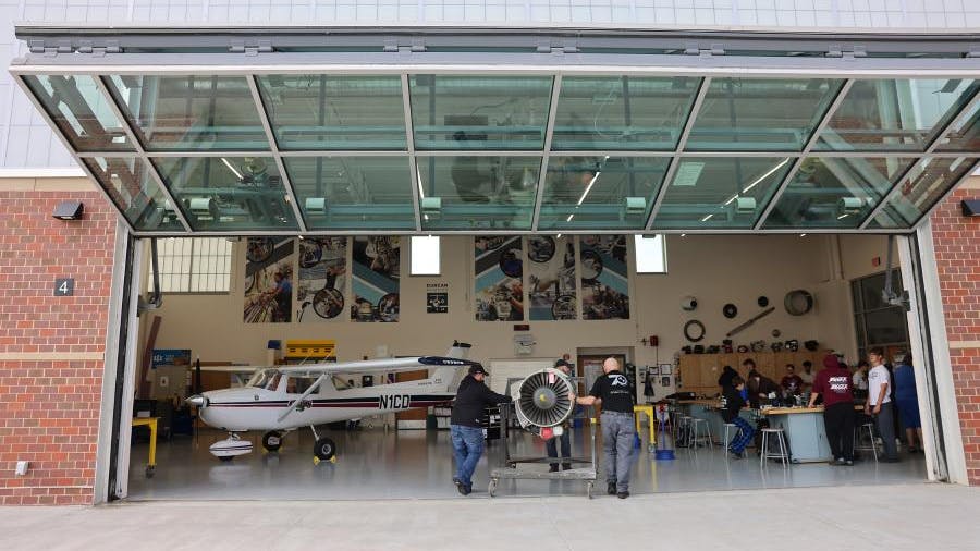 Two people positioning an engine on an engine stand inside a hangar with the door open, with a crowd of people watching to the right