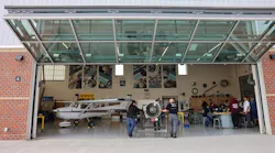 Two people positioning an engine on an engine stand inside a hangar with the door open, with a crowd of people watching to the right Two people positioning an engine on an engine stand inside a hangar with the door open, with a crowd of people watching to the right