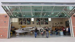 Two people positioning an engine on an engine stand inside a hangar with the door open, with a crowd of people watching to the right Two people positioning an engine on an engine stand inside a hangar with the door open, with a crowd of people watching to the right