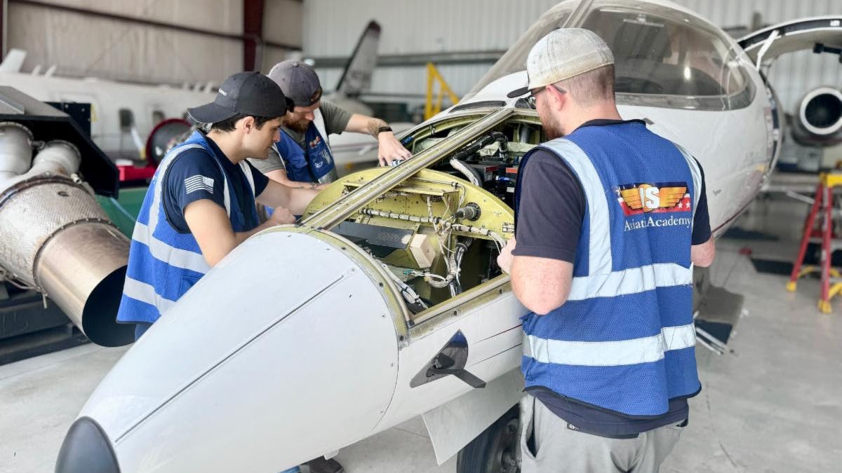 Three aviation maintenance technicians wearing blue vests working on an opened-up airplane inside a hangar