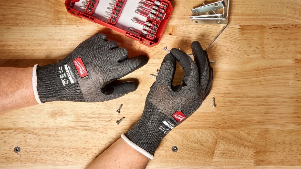 A pair of hands wearing black gloves and holding a screw over a wooden table