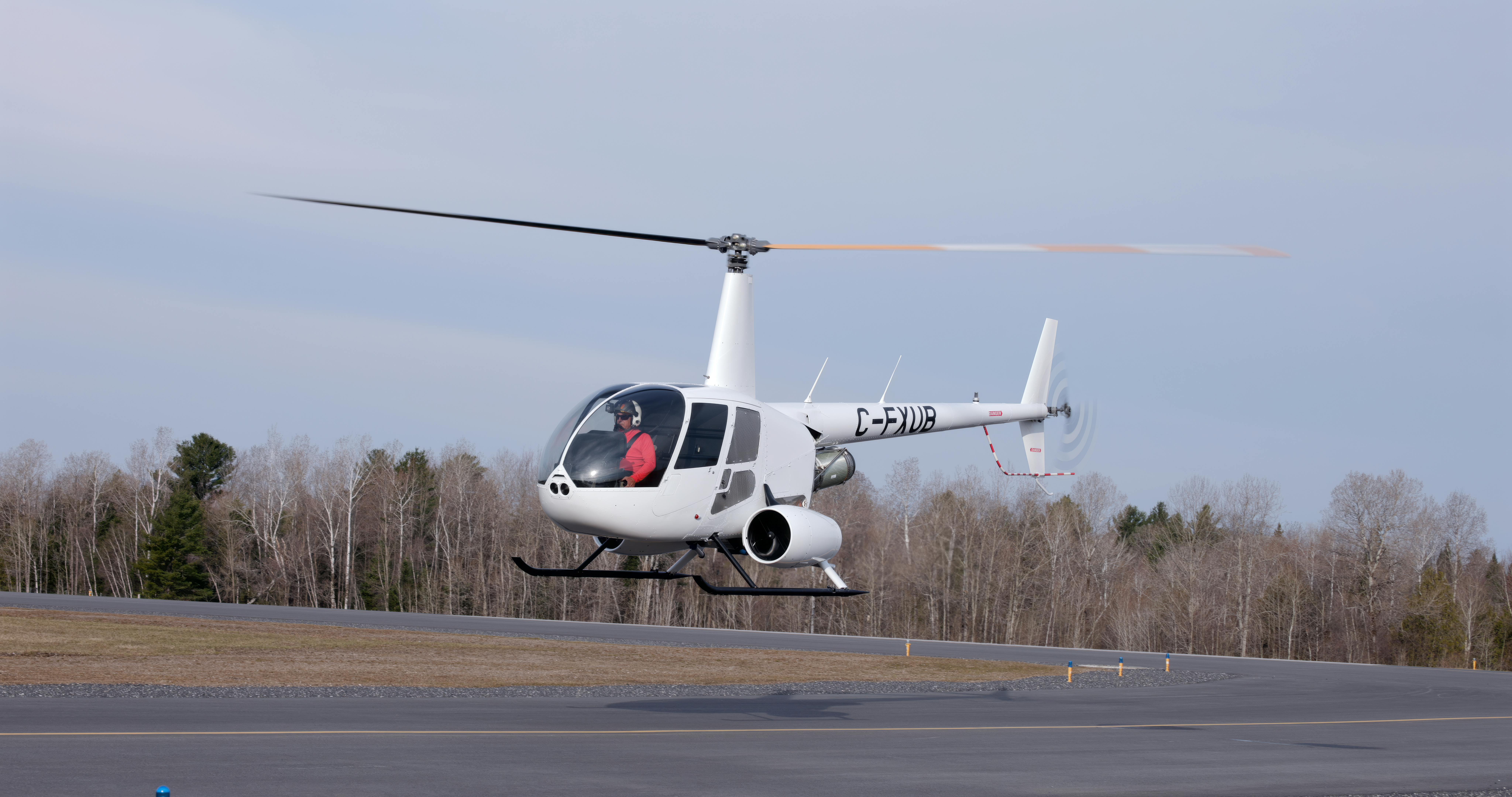 A white helicopter flying above a runway with a pilot in the cockpit and trees in the background