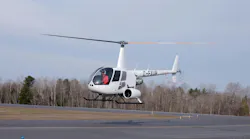 A white helicopter flying above a runway with a pilot in the cockpit and trees in the background