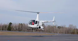 A white helicopter flying above a runway with a pilot in the cockpit and trees in the background A white helicopter flying above a runway with a pilot in the cockpit and trees in the background