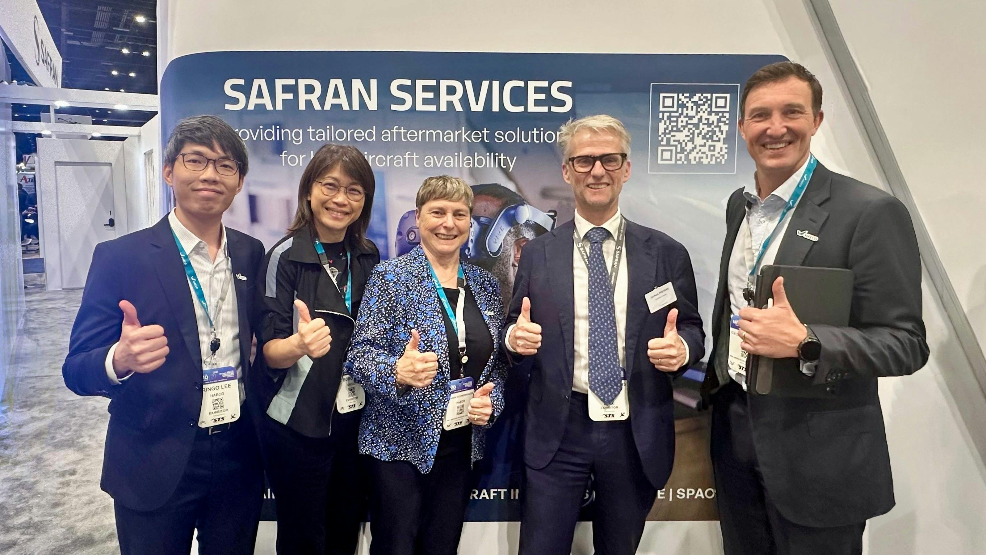 A group of men and women wearing business attire and ID badges, smiling and giving a thumbs-up to the camera while standing in front of a sign that reads, 'Safran Services'