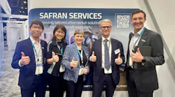 A group of men and women wearing business attire and ID badges, smiling and giving a thumbs-up to the camera while standing in front of a sign that reads, 'Safran Services'