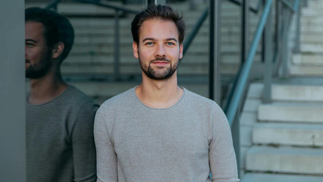 A man wearing a grey shirt leaning against a glass wall and smiling at the camera