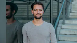 A man wearing a grey shirt leaning against a glass wall and smiling at the camera A man wearing a grey shirt leaning against a glass wall and smiling at the camera