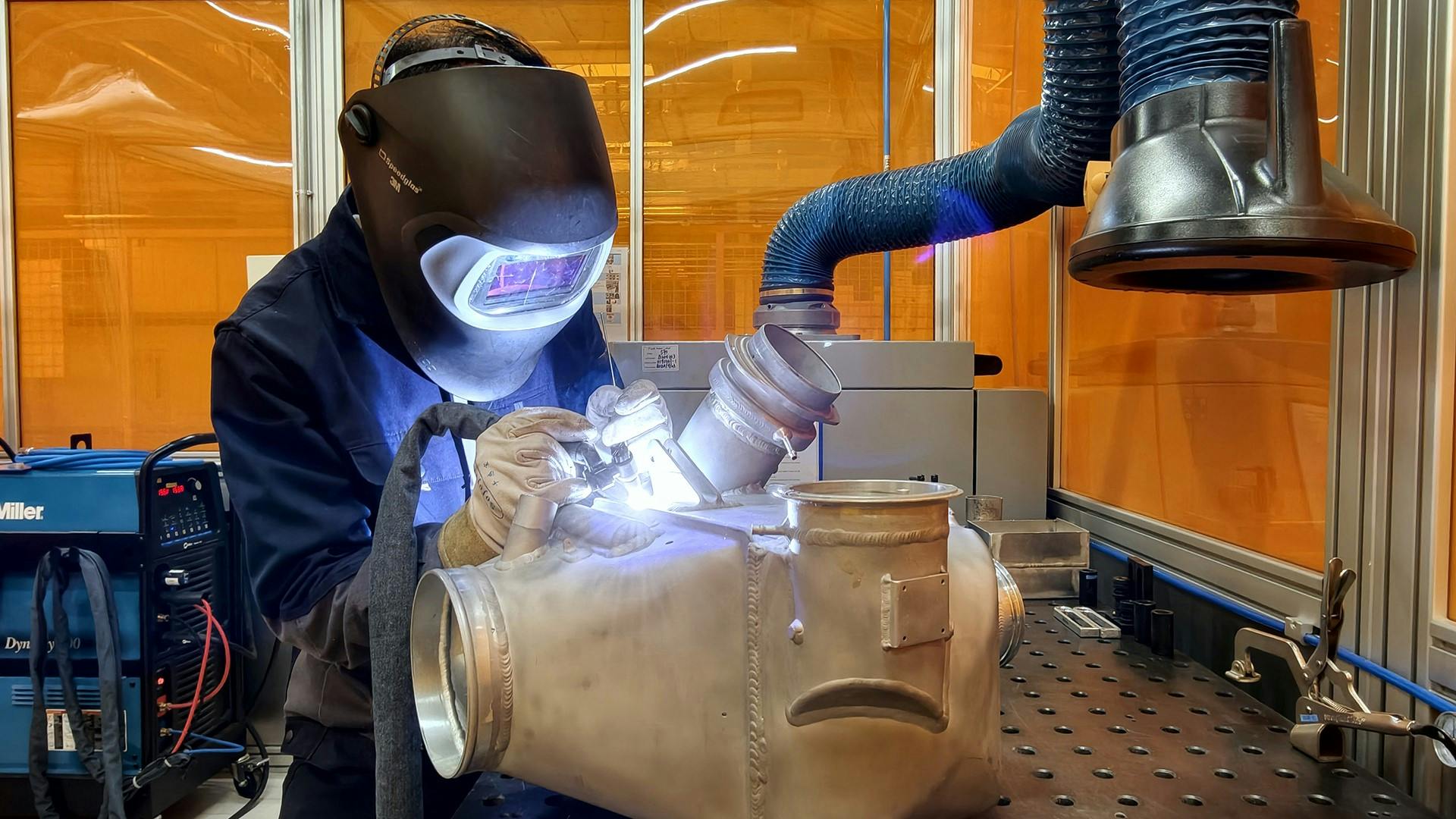A technician wearing heavy PPE including a metal face shield and gloves working on a metal component with a hand tool