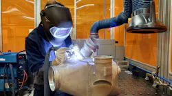 A technician wearing heavy PPE including a metal face shield and gloves working on a metal component with a hand tool