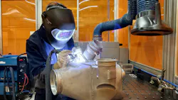 A technician wearing heavy PPE including a metal face shield and gloves working on a metal component with a hand tool A technician wearing heavy PPE including a metal face shield and gloves working on a metal component with a hand tool