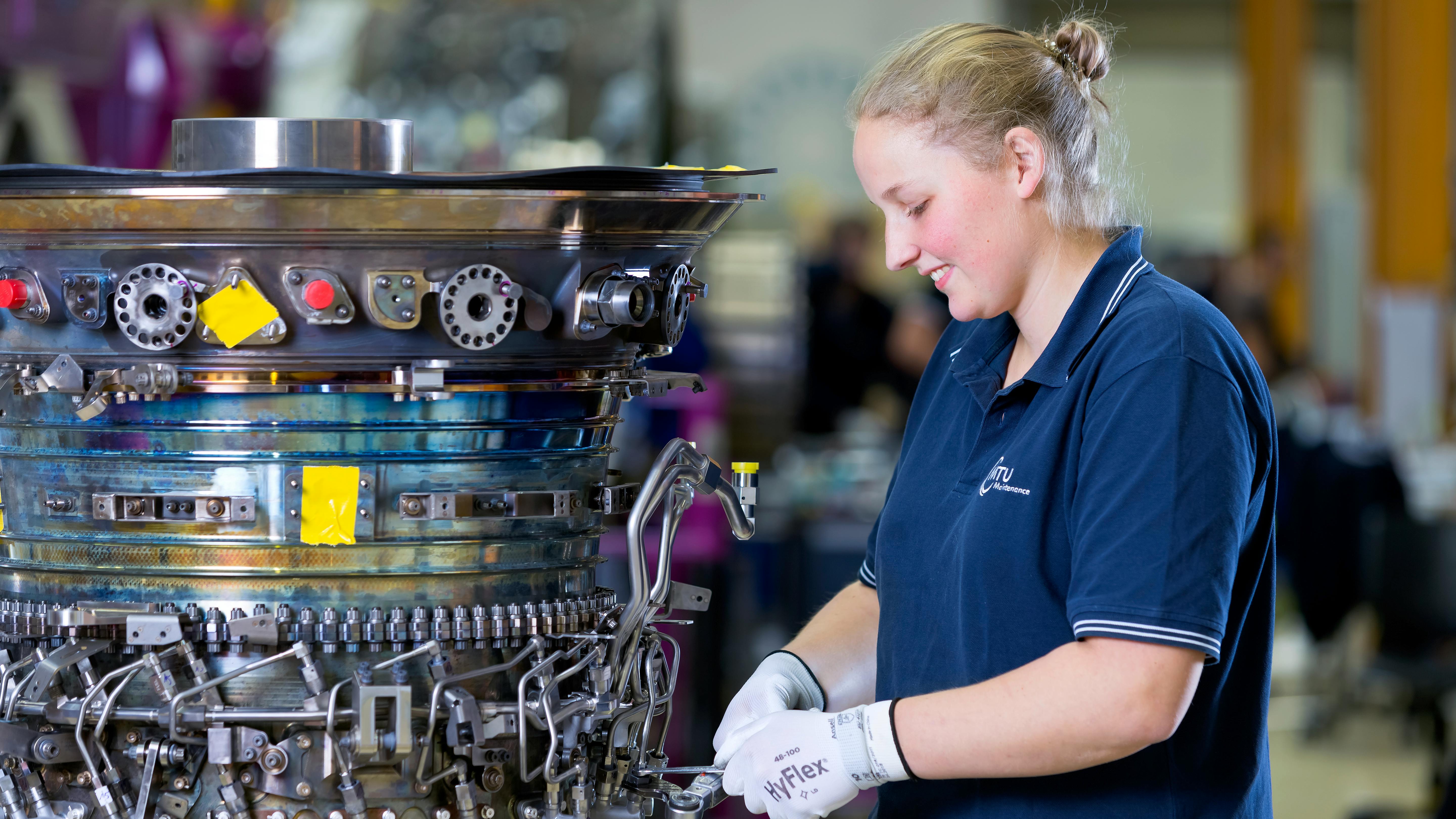 A woman wearing a blue shirt and white gloves working on an aircraft component that's removed from the airplane