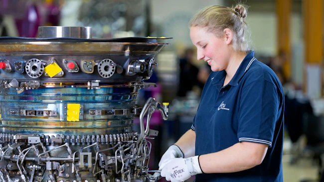 A woman wearing a blue shirt and white gloves working on an aircraft component that's removed from the airplane