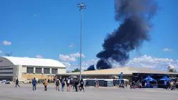 A scattered crowd of people at an outdoor event with black plumes of smoke in the background A scattered crowd of people at an outdoor event with black plumes of smoke in the background