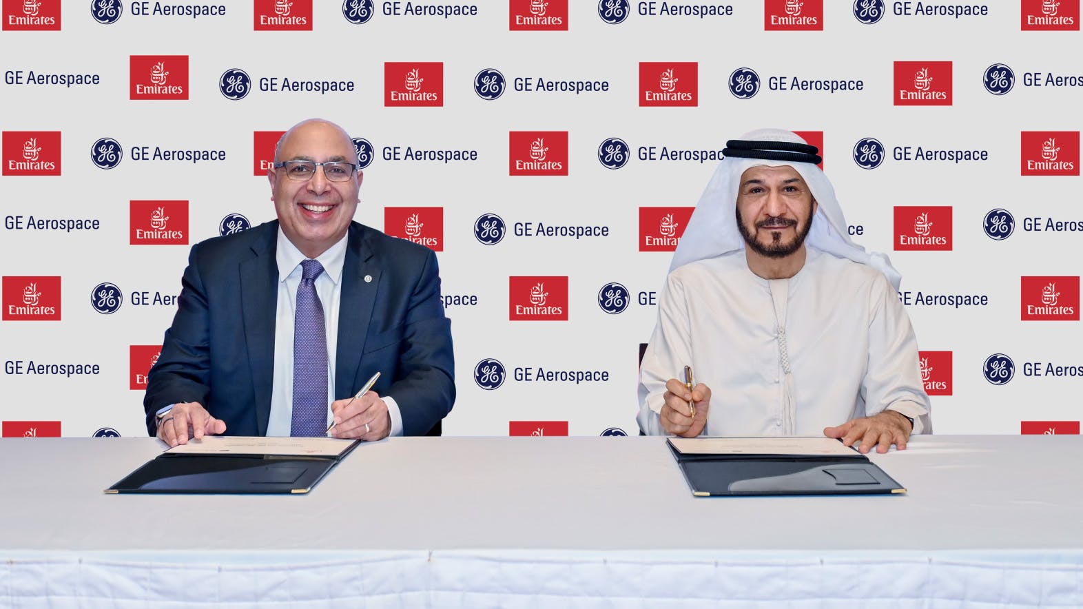 Two men sitting at a table and smiling at the camera while signing documents in front of a backdrop displaying the logos for GE Aerospace and Emirates