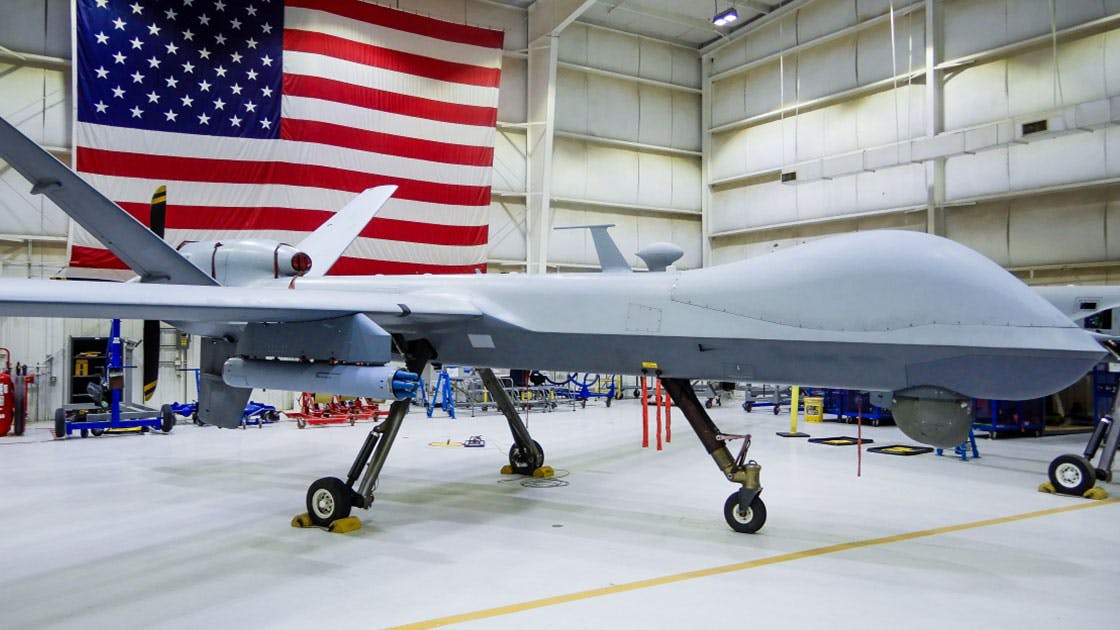 A large white aircraft with no windows parked in a hangar with an American flag hanging in the background