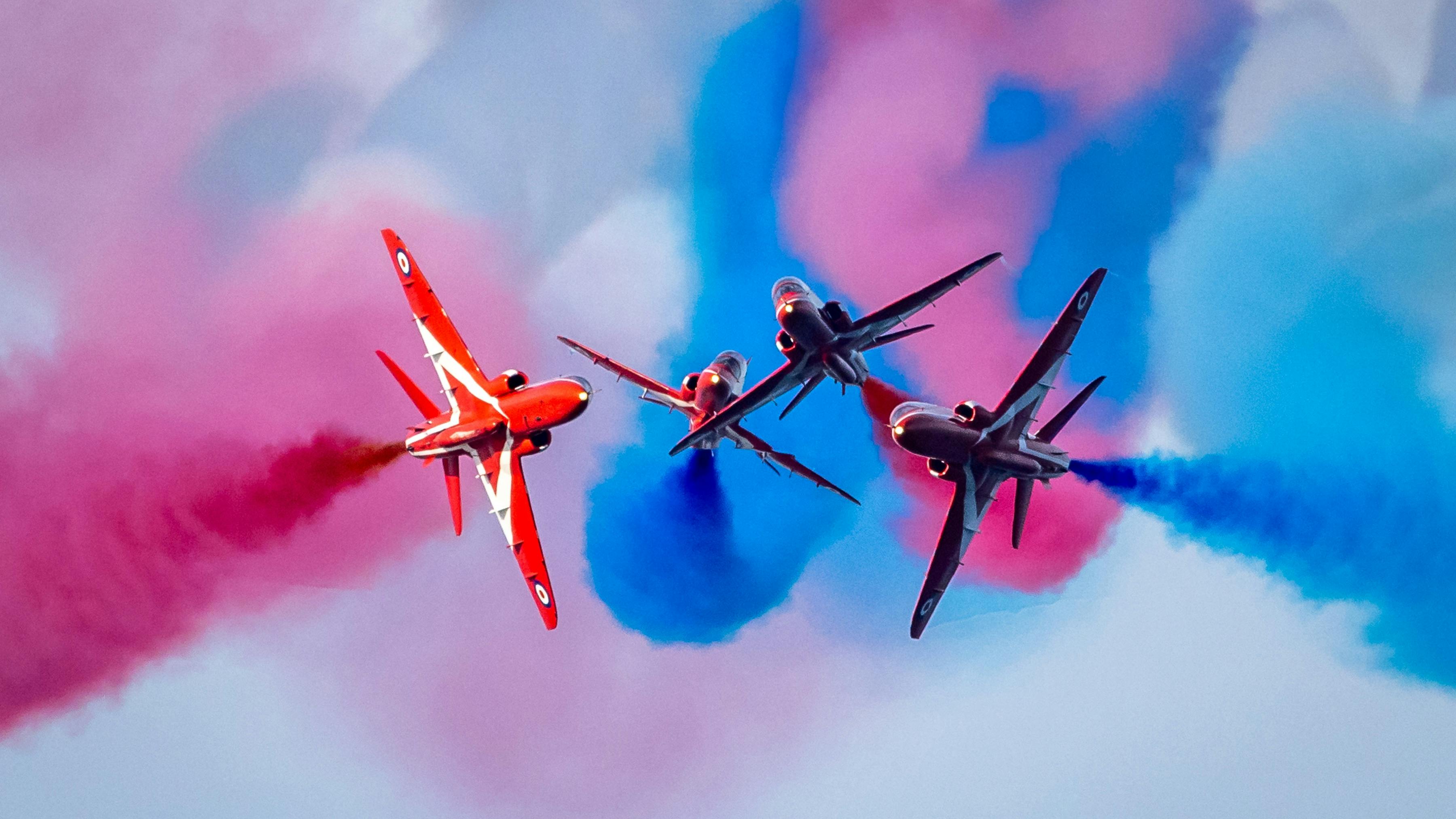 Four red airplanes flying in close formation around each other with pink and blue plumes of smoke in the air