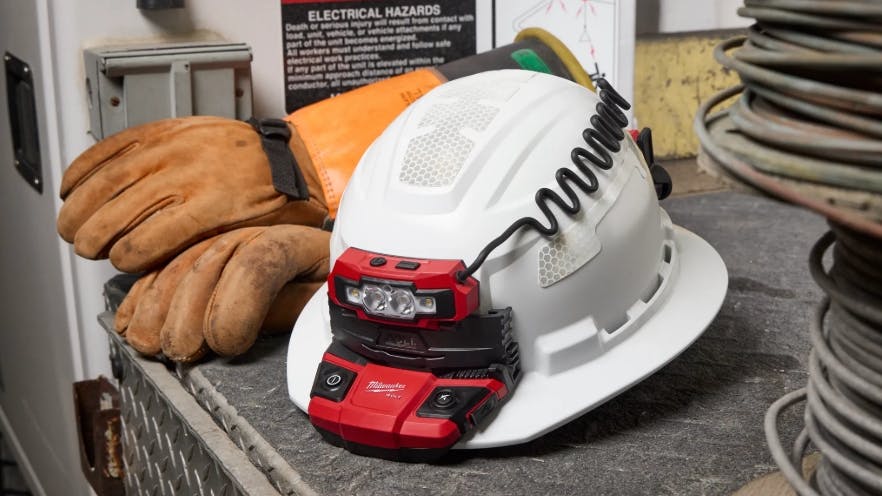 A white hard had with a red attachment on it sitting on a black table with orange gloves sitting behind the helmet