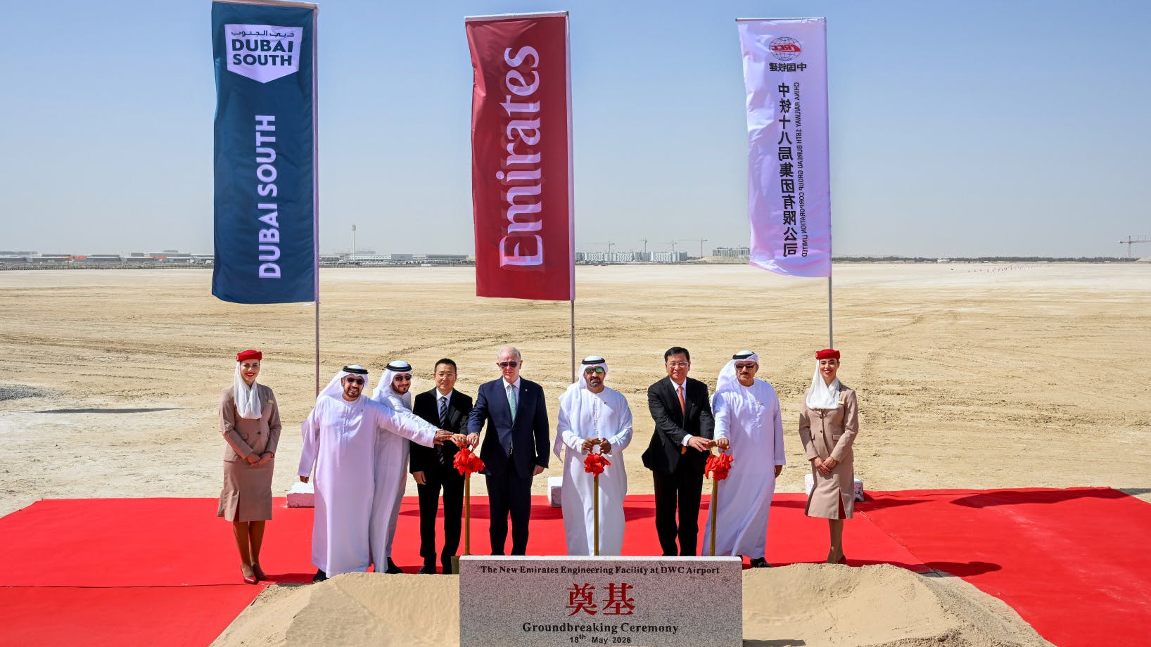 A group of professionals wearing business attire posing with shovels at a construction site for a groundbreaking ceremony, with flags for Emirates, Dubai South and China Railway Construction Corporation in the background