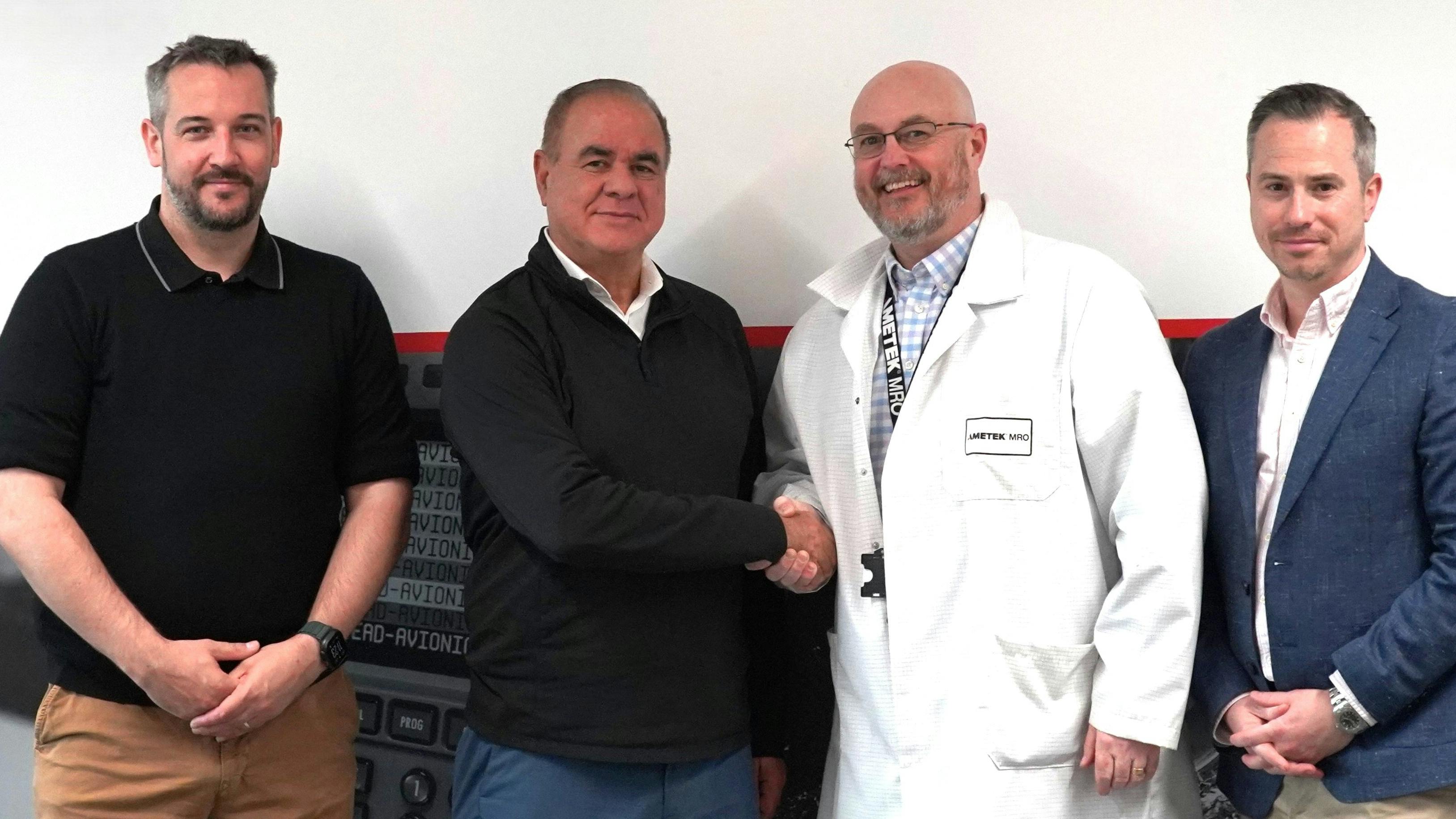 Four men standing in a line and smiling at the camera, with three dressed in business-casual attire and one in a white lab coat