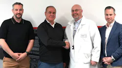 Four men standing in a line and smiling at the camera, with three dressed in business-casual attire and one in a white lab coat