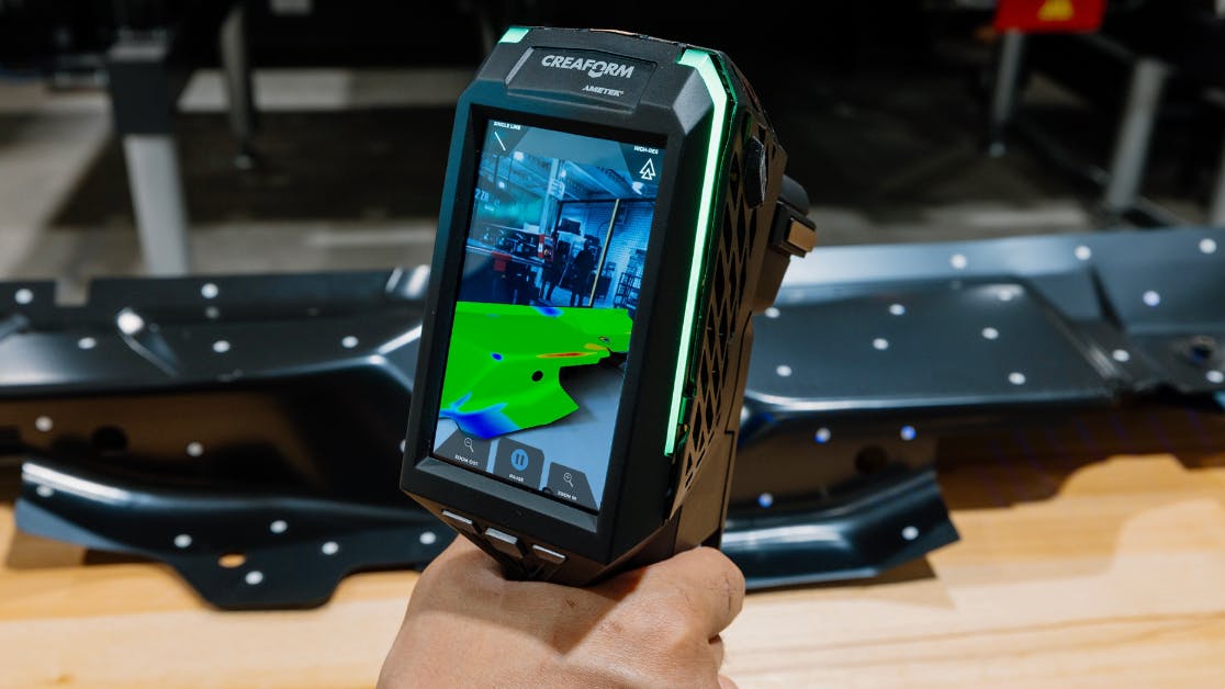 A handheld device with a screen showing a rendering of a piece of metal that's sitting on the ground a few feet away