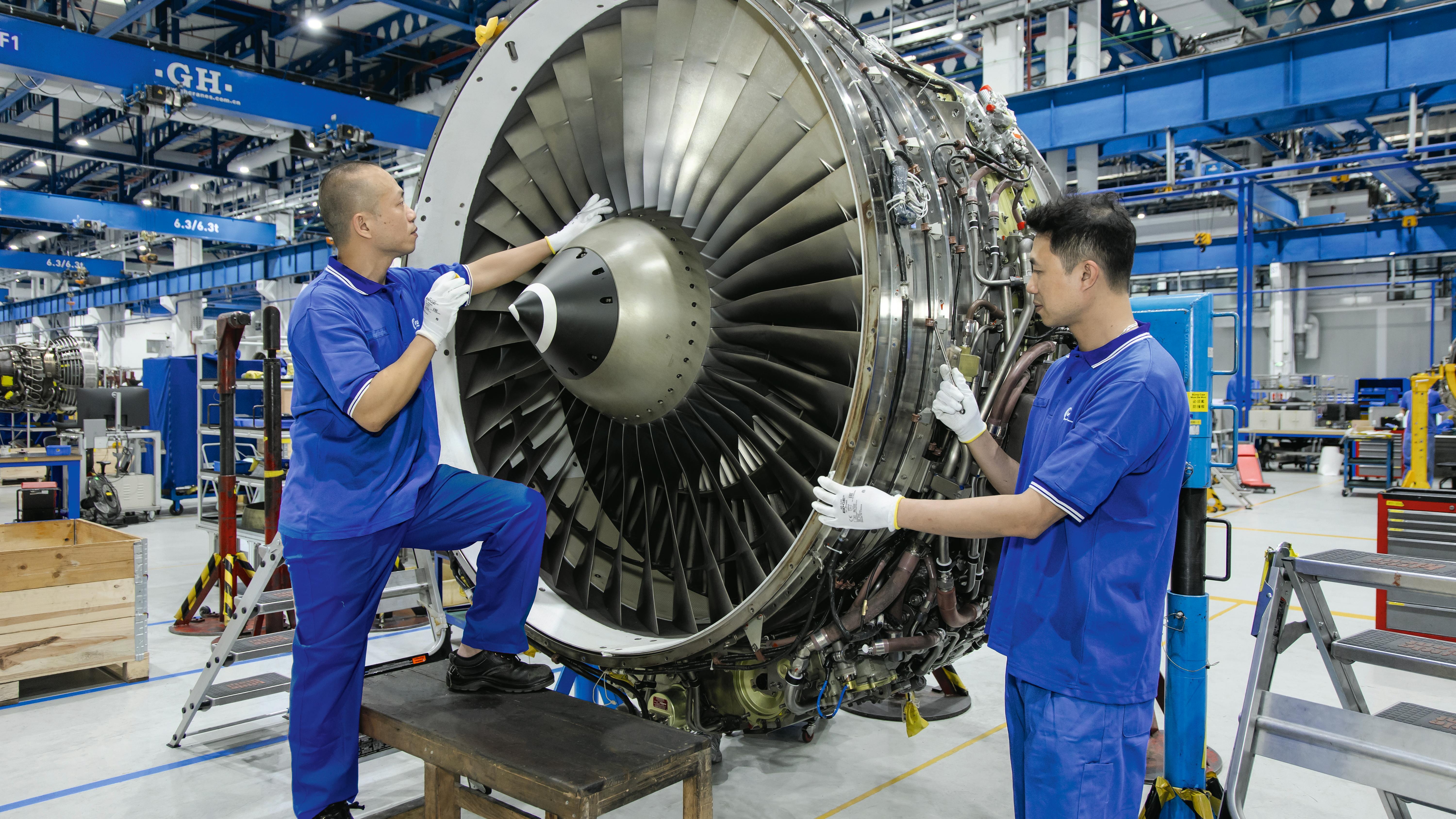 Two technicians wearing blue uniforms working on an airplane engine that's detached from a plane