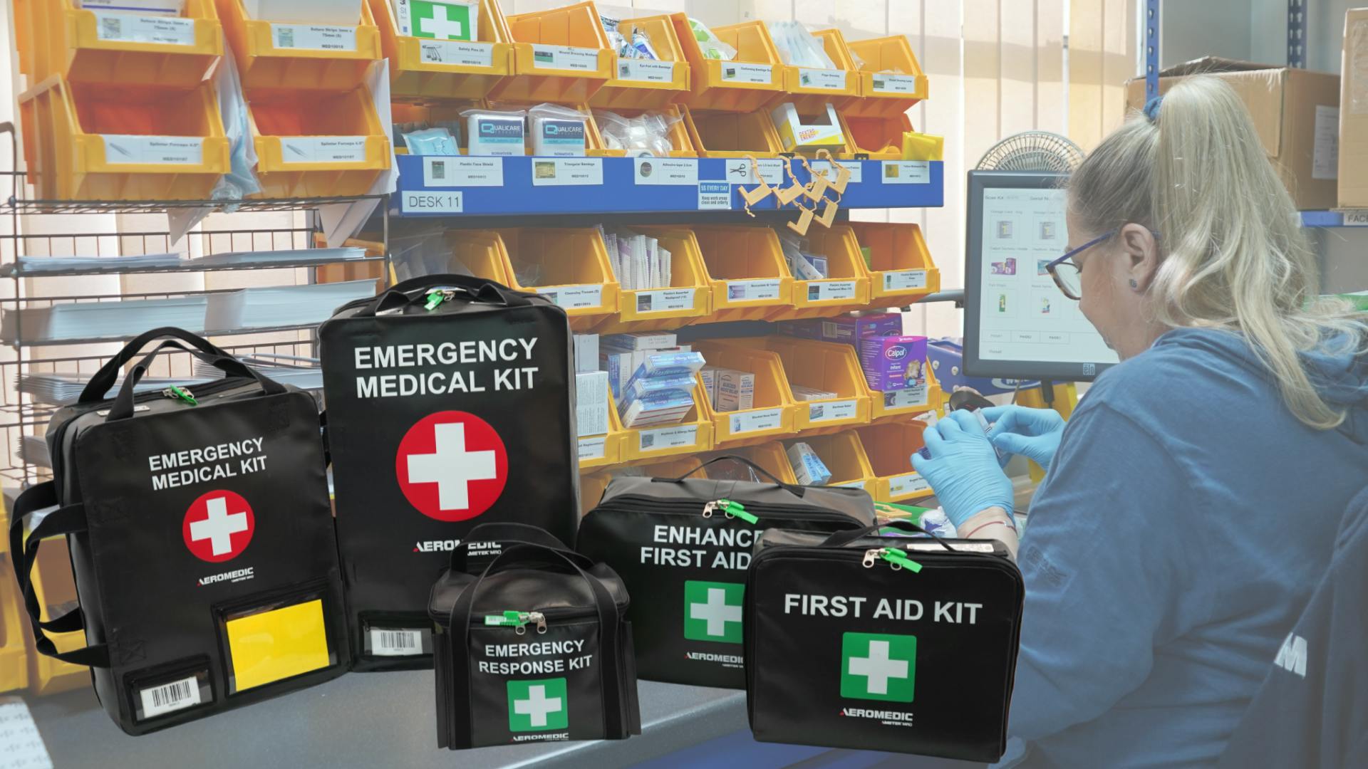 A woman wearing a blue hoodie and blue rubber gloves working next to a group of soft-case medical kits with labels like 'Emergency Medical Kit' and 'First Aid Kit'