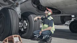 A technician wearing PPE crouching under an airplane near its landing gear A technician wearing PPE crouching under an airplane near its landing gear