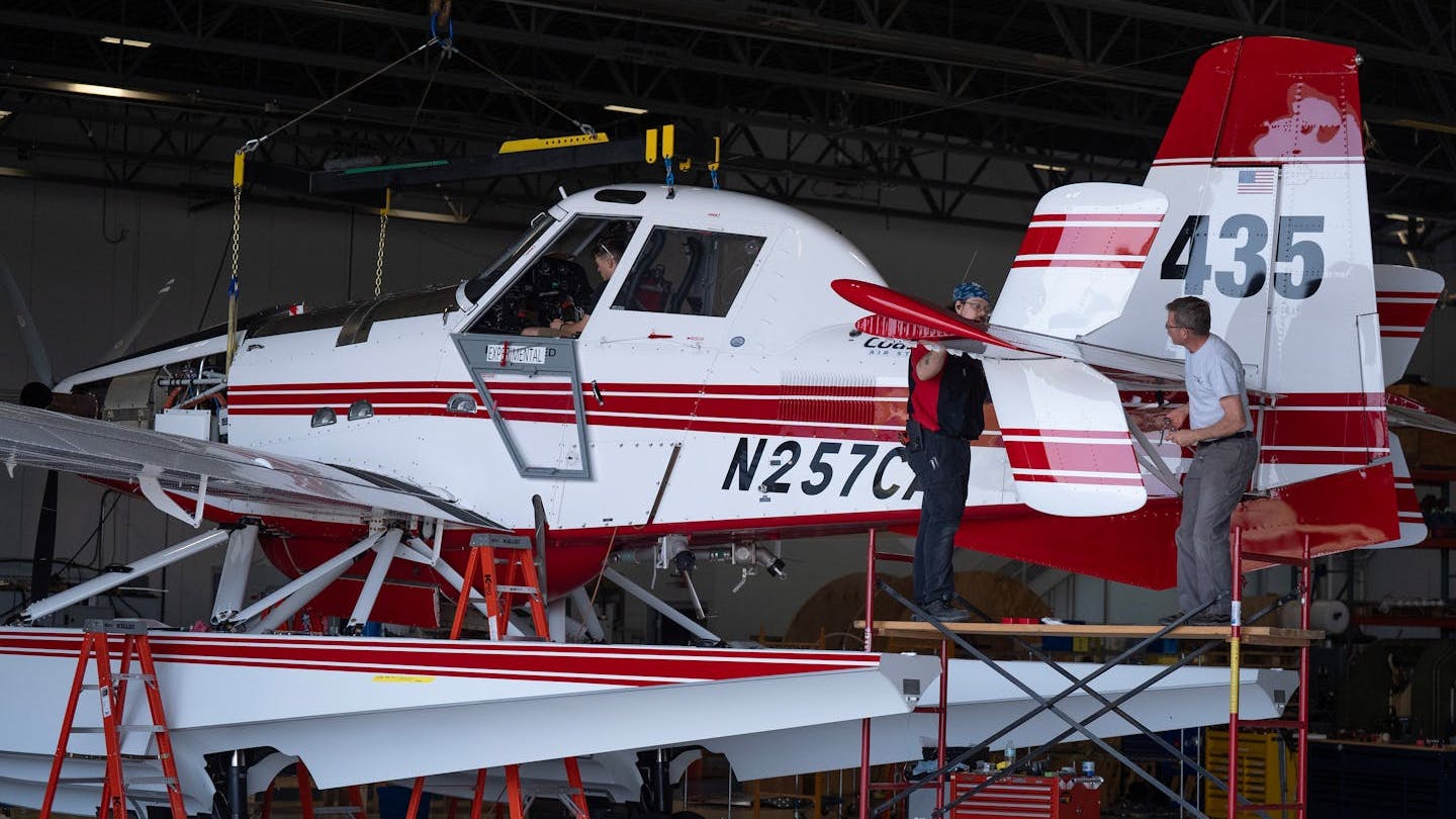 A red and white seaplane parked on a platform while a man checks a rear compartment