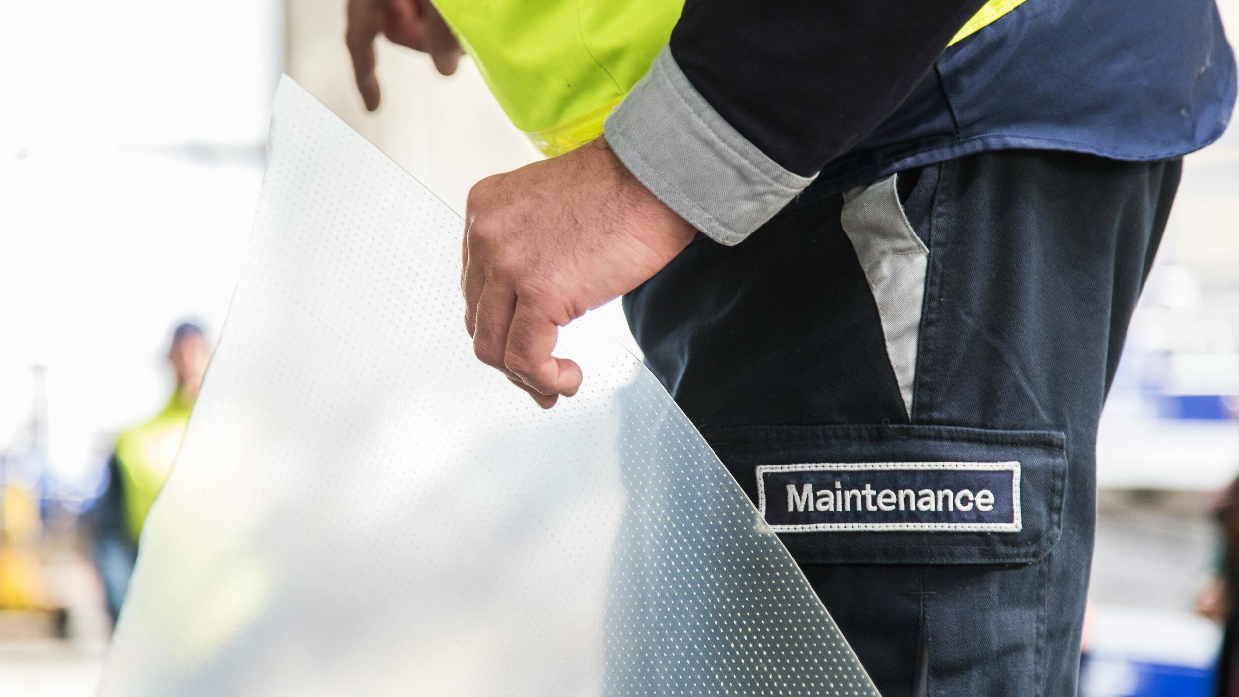 A technician wearing pants with a patch that says, 'MAINTENANCE' holding a piece of clear film with riblets on it