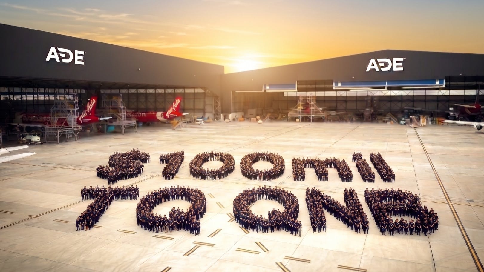 A wide shot of an aircraft hangar with 'ADE' written on the buildings and a large crowd of people gathered outside in a formation displaying the following text: $100 MIL TQ QNB