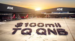 A wide shot of an aircraft hangar with 'ADE' written on the buildings and a large crowd of people gathered outside in a formation displaying the following text: $100 MIL TQ QNB