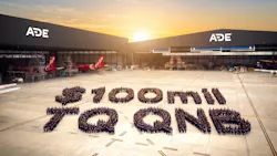 A wide shot of an aircraft hangar with 'ADE' written on the buildings and a large crowd of people gathered outside in a formation displaying the following text: $100 MIL TQ QNB A wide shot of an aircraft hangar with 'ADE' written on the buildings and a large crowd of people gathered outside in a formation displaying the following text: $100 MIL TQ QNB