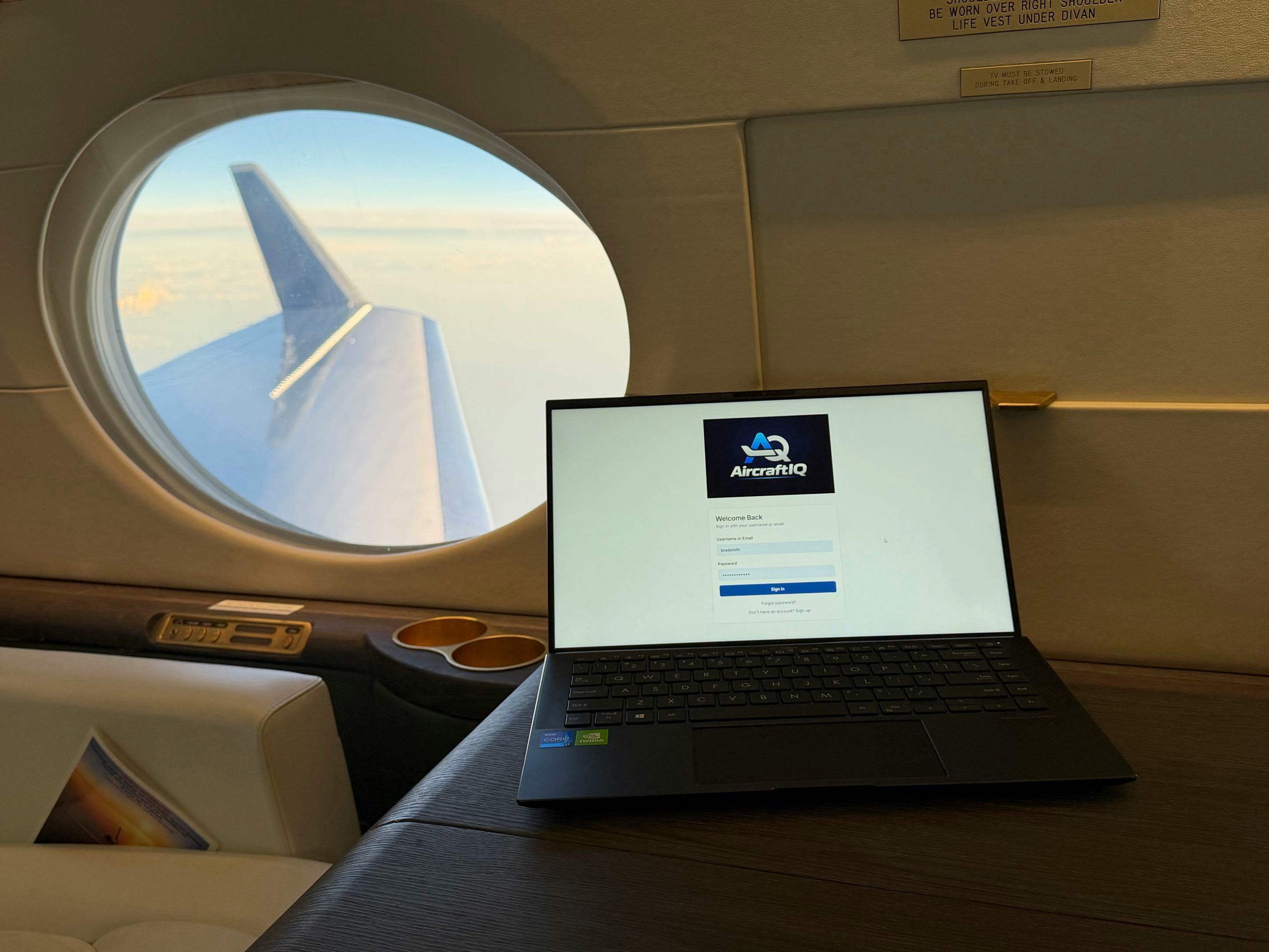 A laptop perched on someone's lap displaying a homepage with text that says. 'AircraftIQ' while seated on an airplane in the air, the window in the background