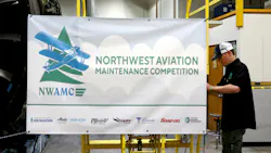 A young man putting up a banner that reads: NWAMC Northwest Aviation Maintenance Competition A young man putting up a banner that reads: NWAMC Northwest Aviation Maintenance Competition