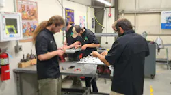 Three young men working in a mechanical workshop gathered around a gray work table Three young men working in a mechanical workshop gathered around a gray work table