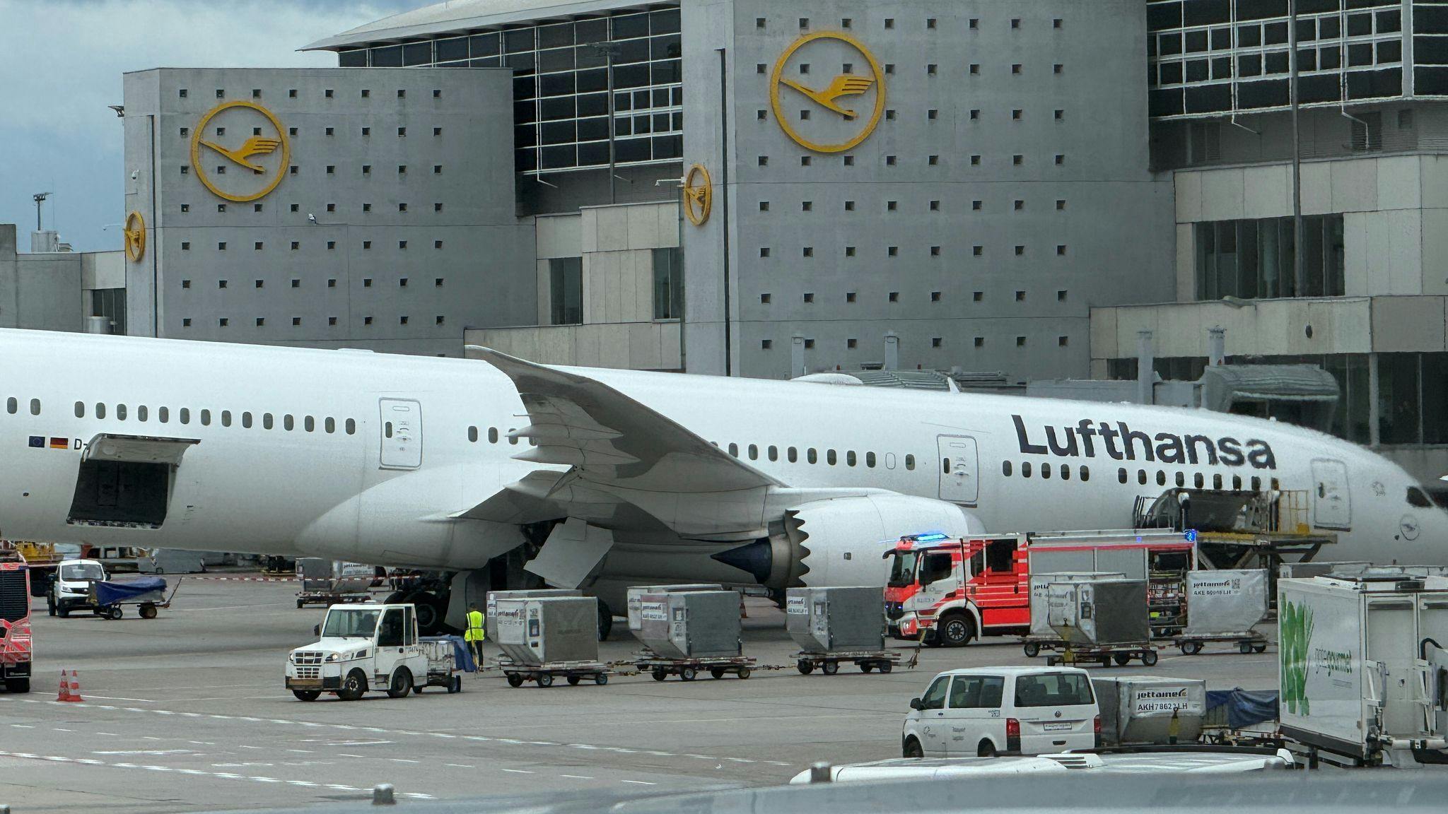 An airplane on the ground with ground handling equipment driving around it, with the word 'Lufthansa' printed on the plane