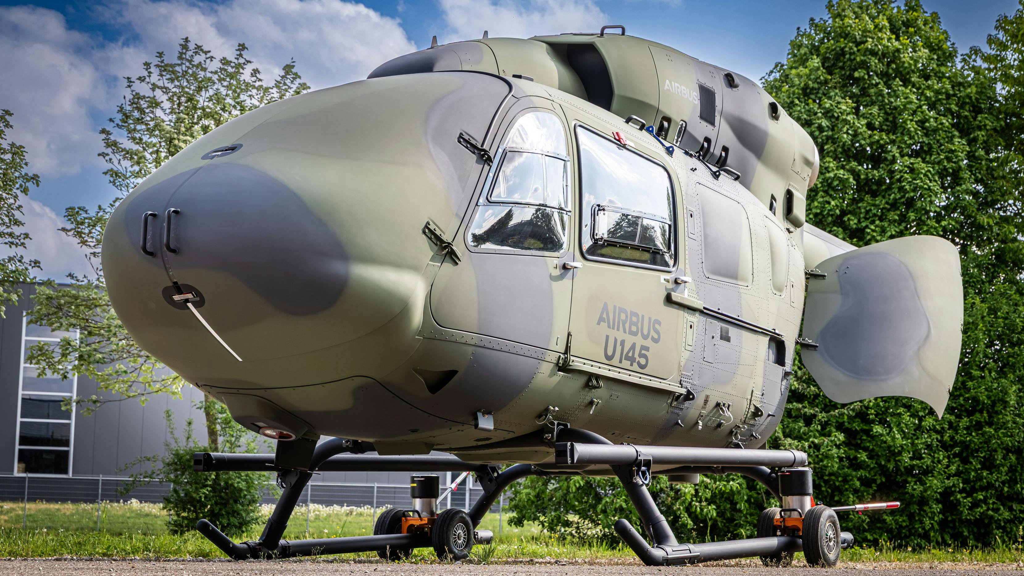 A military aircraft with a round shape and camouflage coloring parked on the ground outside near trees and a building