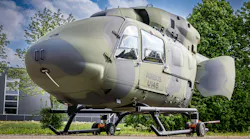 A military aircraft with a round shape and camouflage coloring parked on the ground outside near trees and a building