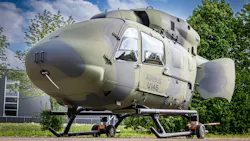 A military aircraft with a round shape and camouflage coloring parked on the ground outside near trees and a building A military aircraft with a round shape and camouflage coloring parked on the ground outside near trees and a building