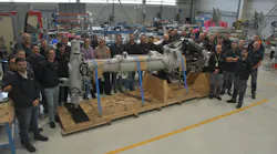 A large group of people gathered around a piece of aircraft landing gear in a repair shop