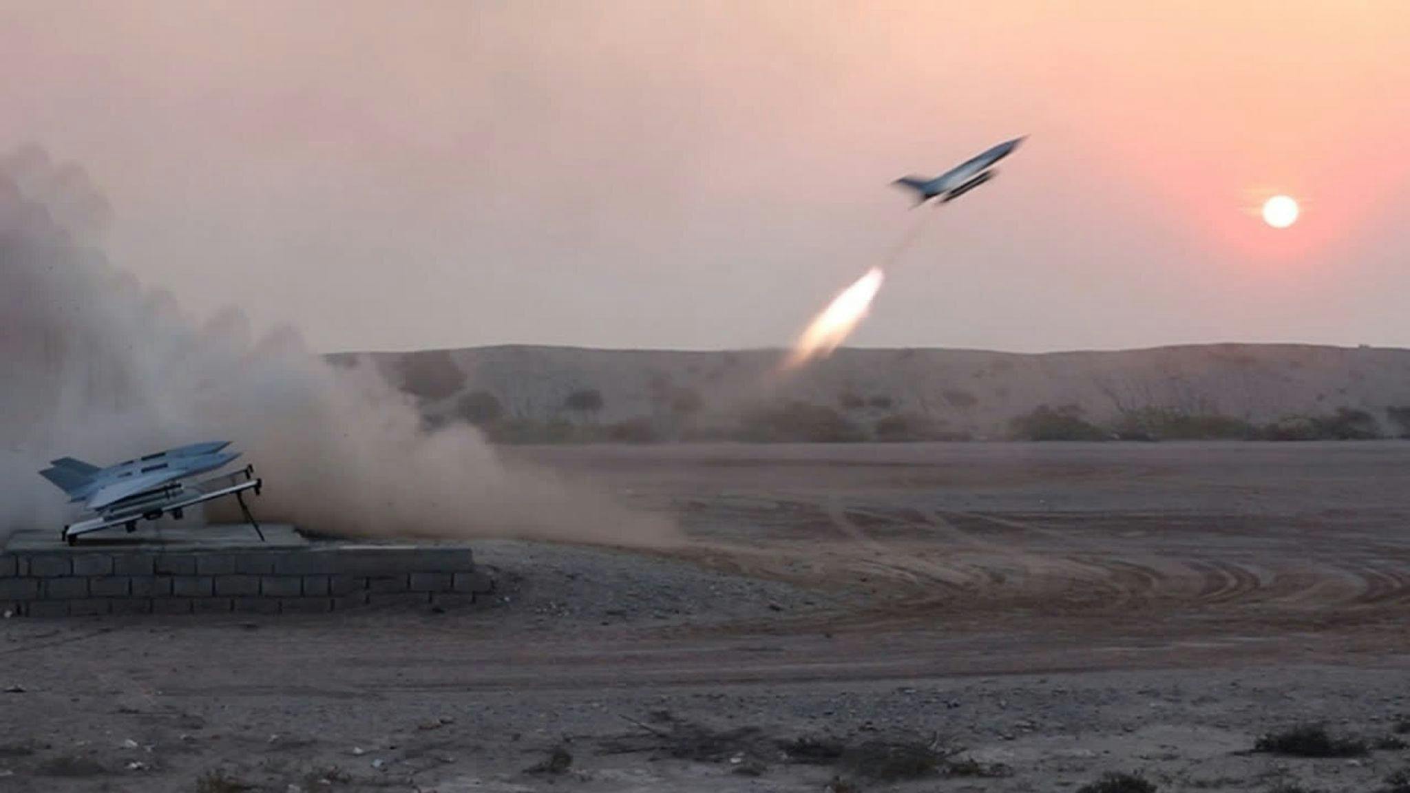 A military aircraft with fire behind it dropping explosives on sand, with clouds of dust rising from the ground