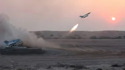 A military aircraft with fire behind it dropping explosives on sand, with clouds of dust rising from the ground A military aircraft with fire behind it dropping explosives on sand, with clouds of dust rising from the ground