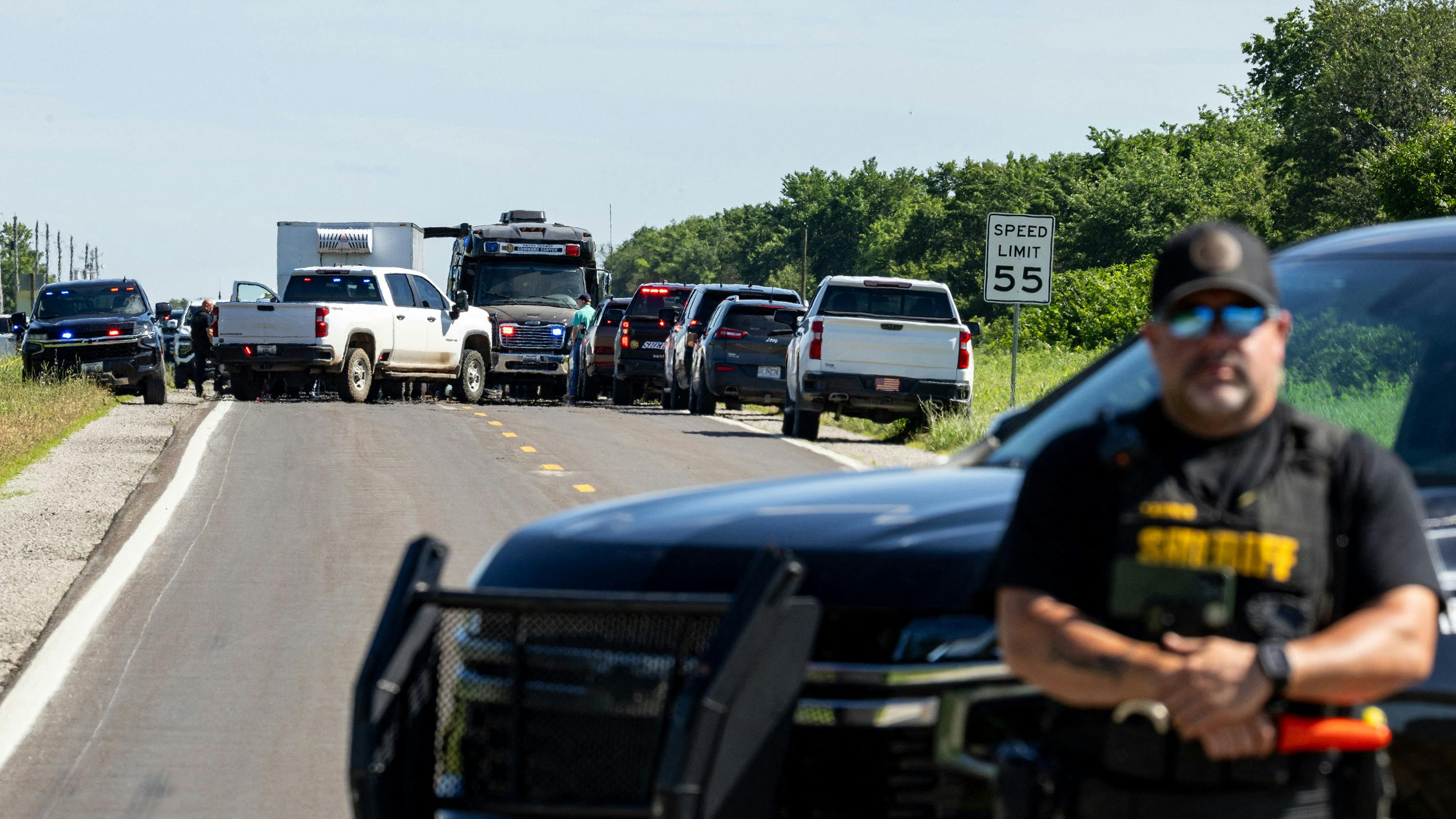 A photograph of a group of vehicles gathered in the road, including emergency vehicles, with a police officer standing in the foreground