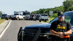 A photograph of a group of vehicles gathered in the road, including emergency vehicles, with a police officer standing in the foreground A photograph of a group of vehicles gathered in the road, including emergency vehicles, with a police officer standing in the foreground