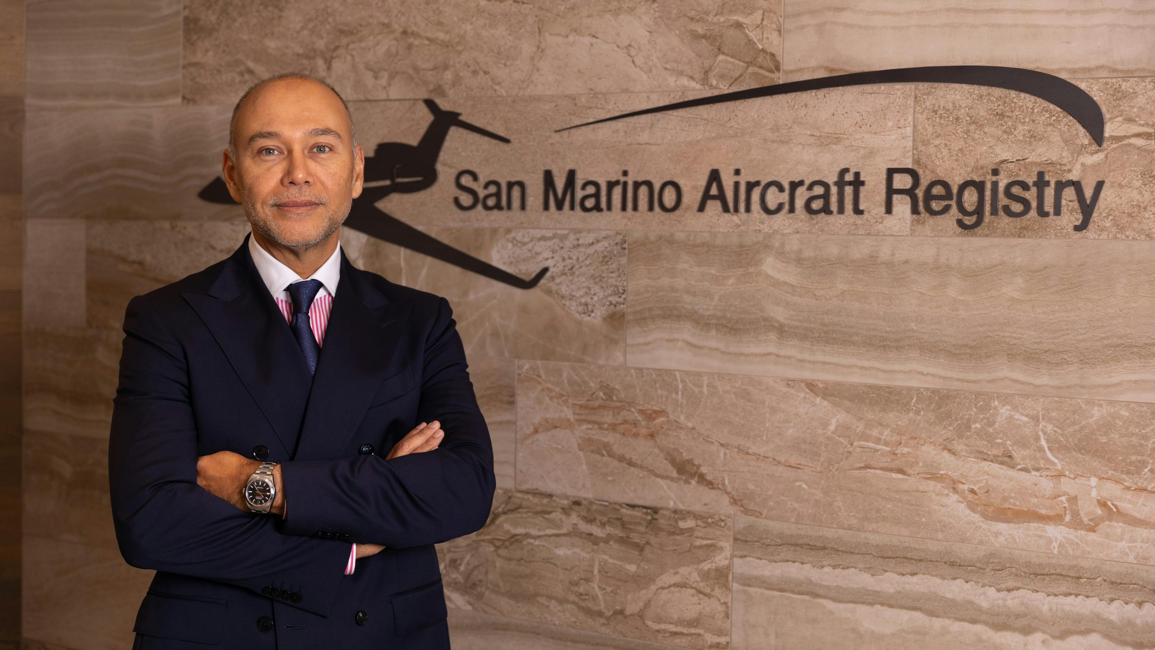 A man wearing a suit crossing his arms and smiling at the camera while standing in front of a wall with a sign that reads: San Marino Aircraft Registry