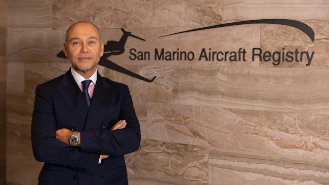 A man wearing a suit crossing his arms and smiling at the camera while standing in front of a wall with a sign that reads: San Marino Aircraft Registry
