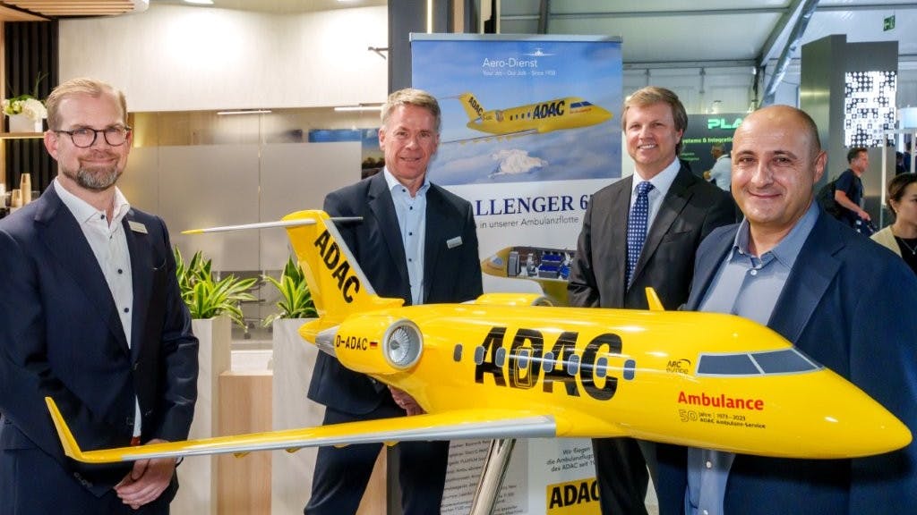 Four men wearing suits smiling at the camera while standing next to a yellow model airplane with text printed on the side that reads: ADAC Ambulance